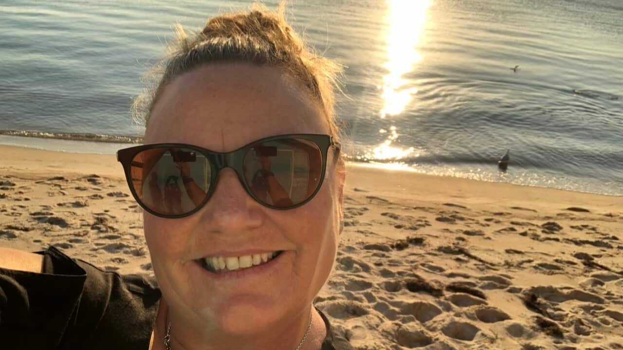 A woman smiles with sunglasses in front of a beach.