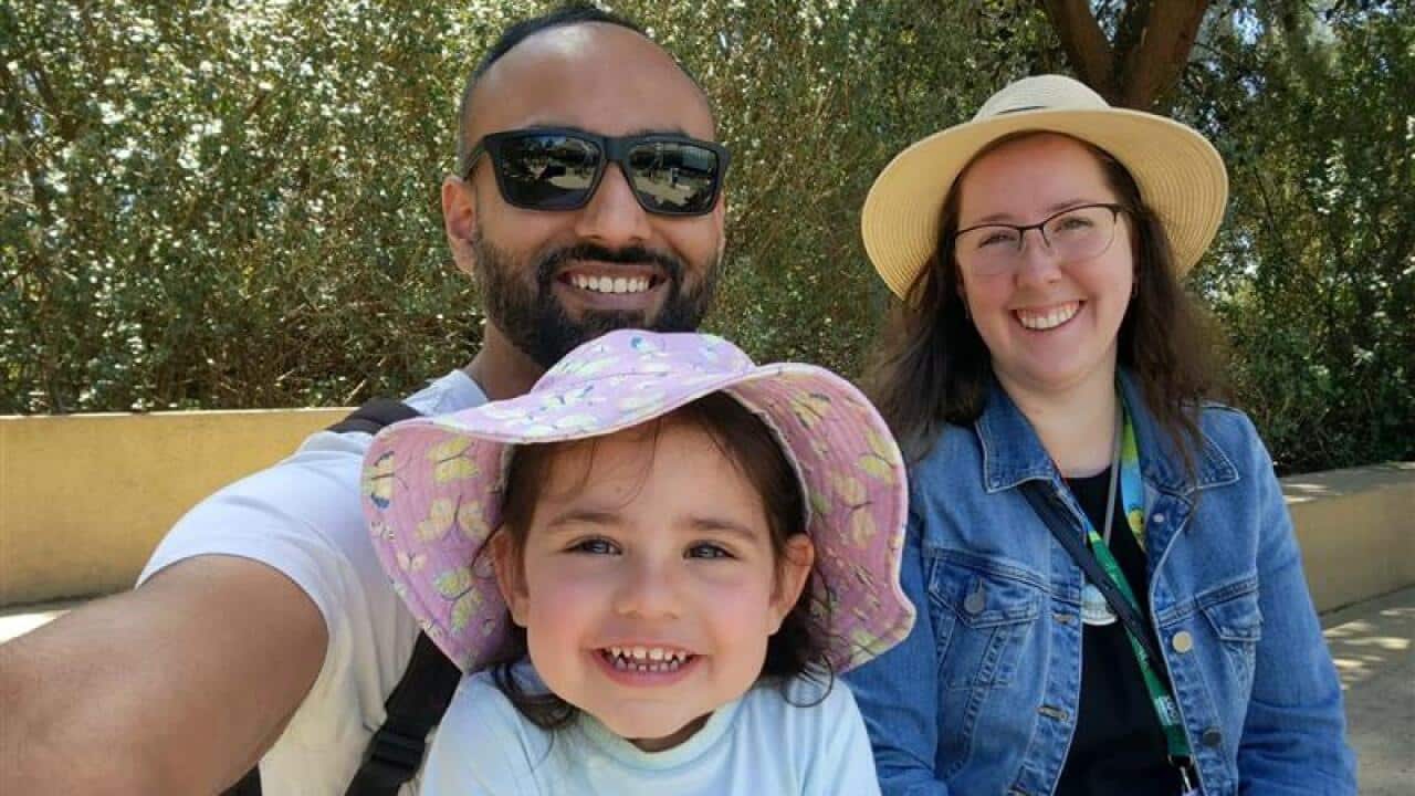 A family of three sit on a bench and smile to camera. The mother and daughter wear hats and the father has sunglasses on.