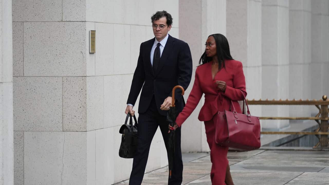 A man wearing a suit walking outside a courtroom, next to a woman wearing a red blazer, red pants and carrying a red bag.