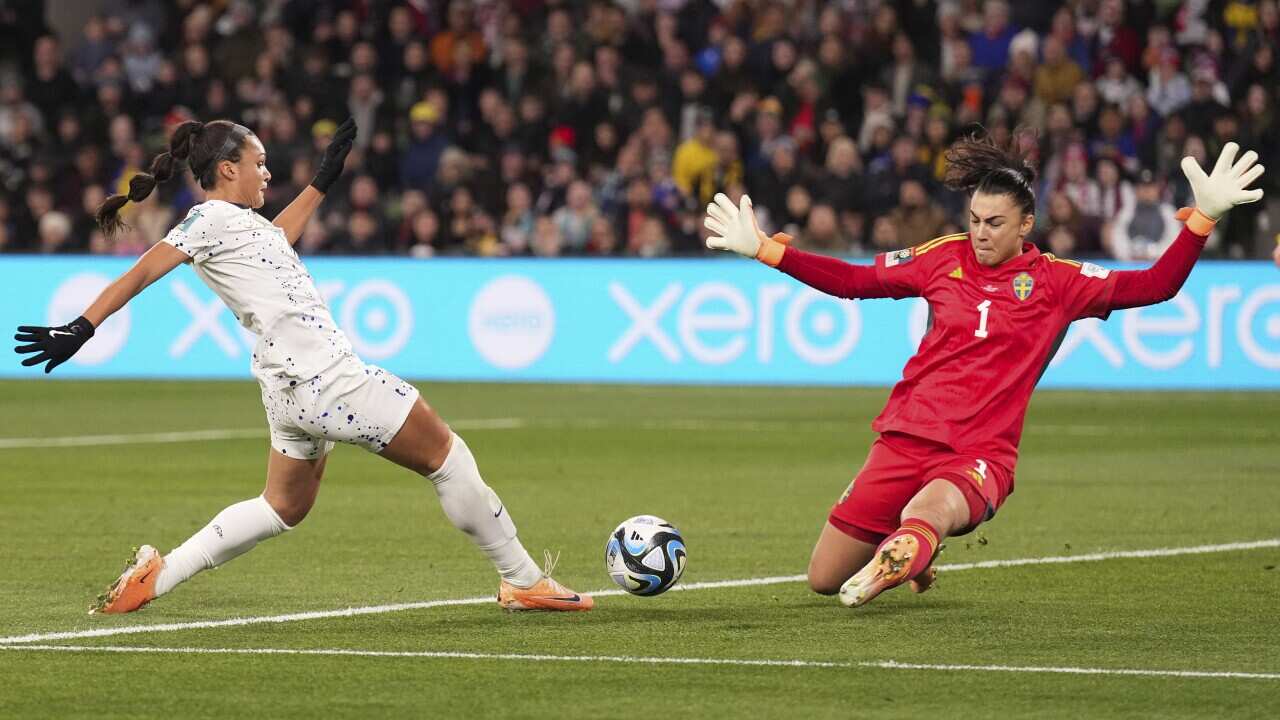 A woman in a white football uniform and a woman in a red football uniform contest a soccer ball.