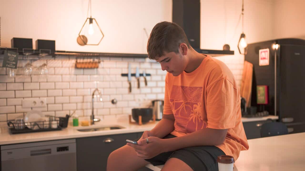 Teenage boy sitting at the kitchen table interacting with his smart phone while having a takeaway coffee