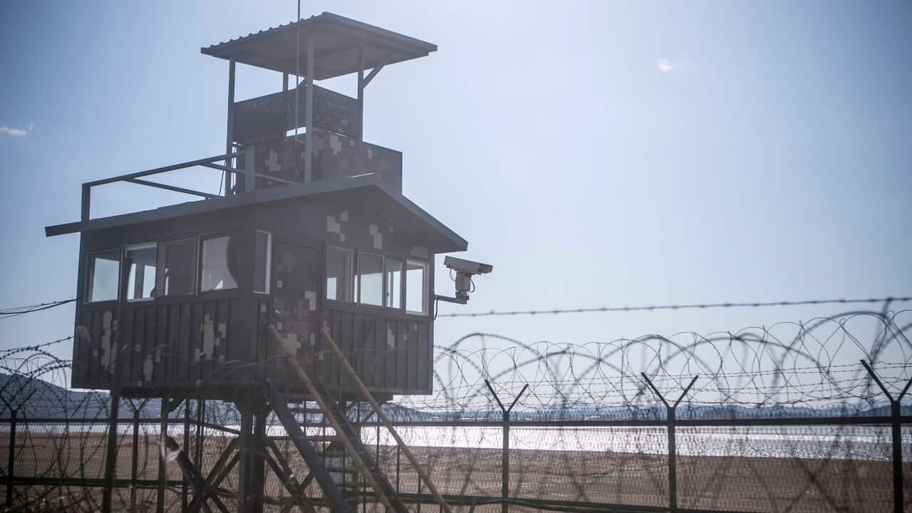 An army post with a large security camera in front of a barbed wire fence.