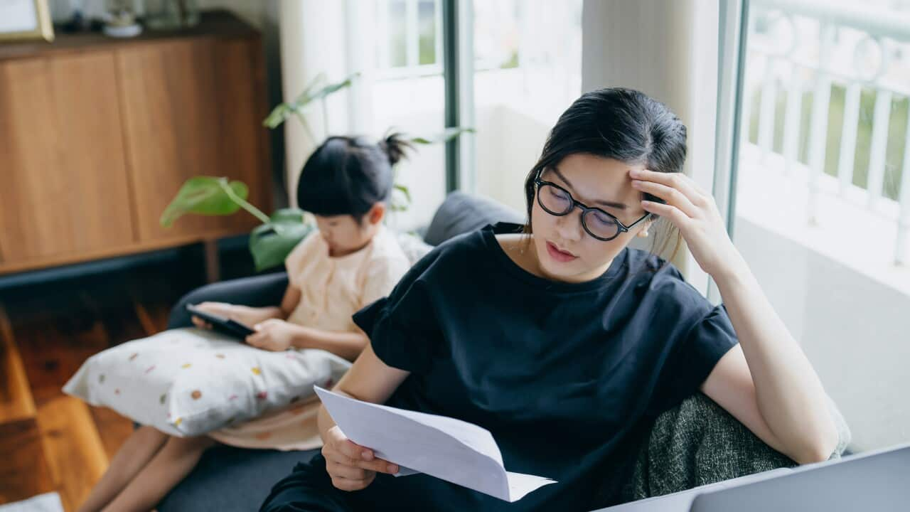 A woman looking stressed while holding paper. A girl is using digital tablet in the background.