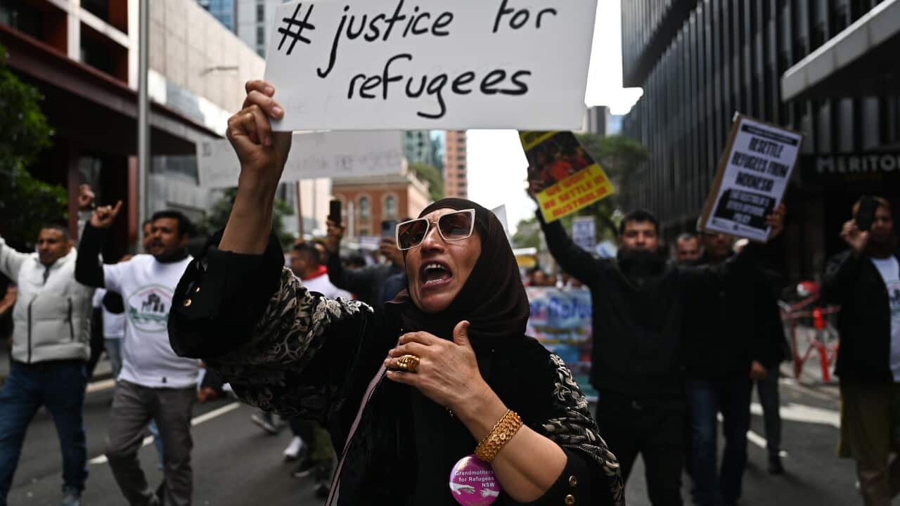 A woman in the foreground holds up a a sign that says #justice for refugees, others on the street behind her are also holding up posters.
