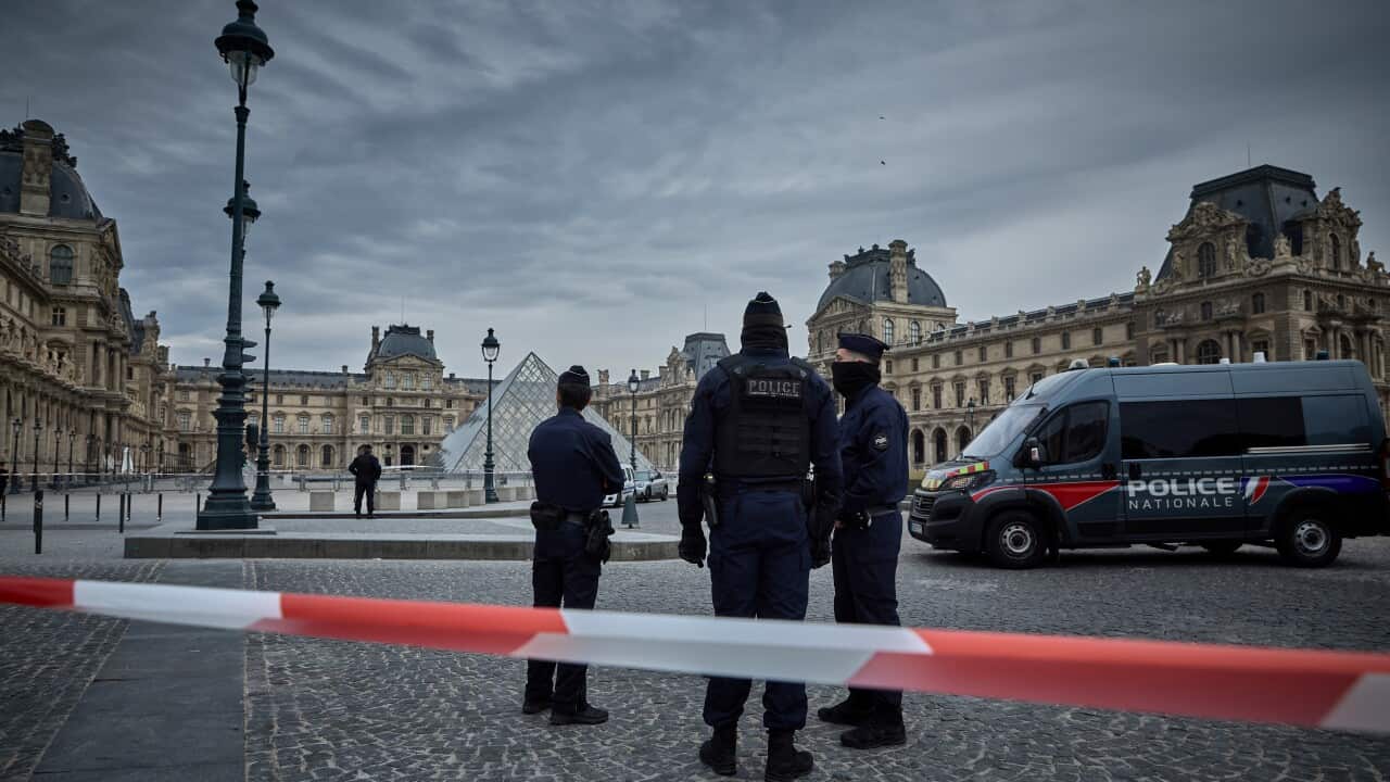 Police officials standing outside a quadrangle of historic buildings, a police vehicle parked nearby and with a police tape in the foreground.