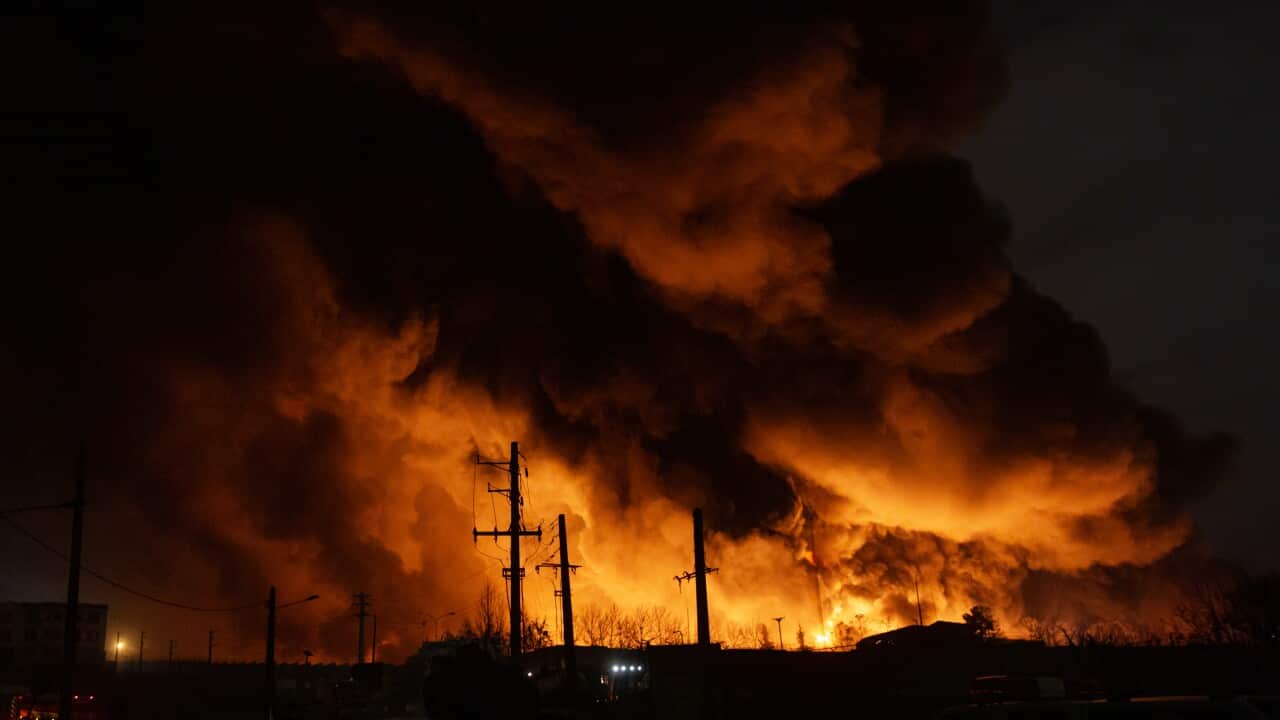 A gigantic fire burns in Shahran oil depot following US and Israeli attacks in Tehran. The photo was taken at night, and the flames are a bright orange with vast plumes of billowing black smoke above them. In the foreground, silhouetted against the flames, are a series of telephone poles, some of their wires already melted.