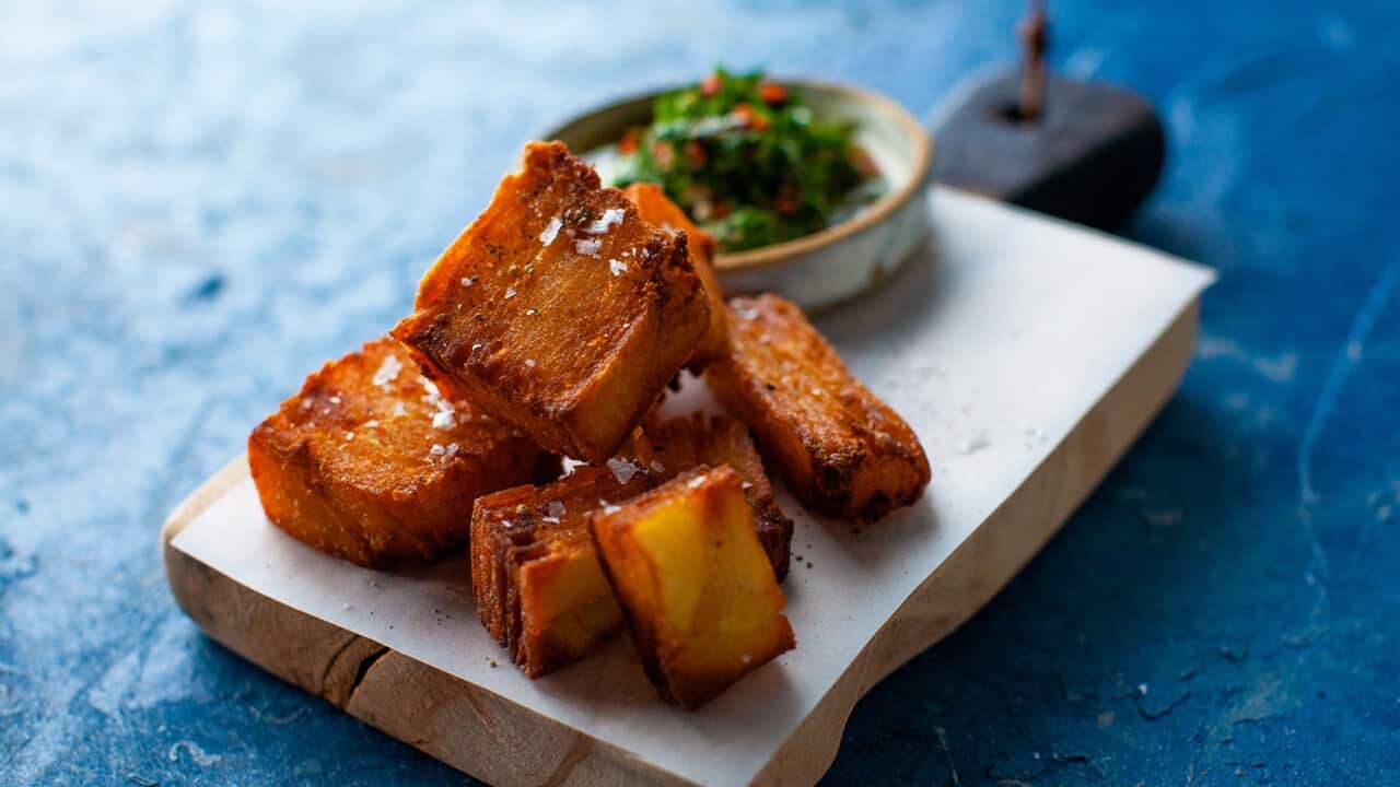 A pile of crunchy, dark golden brown potato chunks sit on a board, along with a bowl of green sauce.