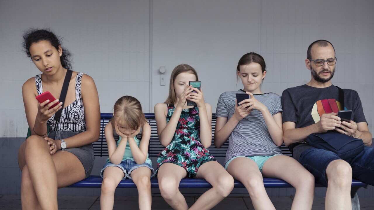 Adults and children sitting on a bench distracted by mobile phones whilst younger child without a phone is completely fed up
