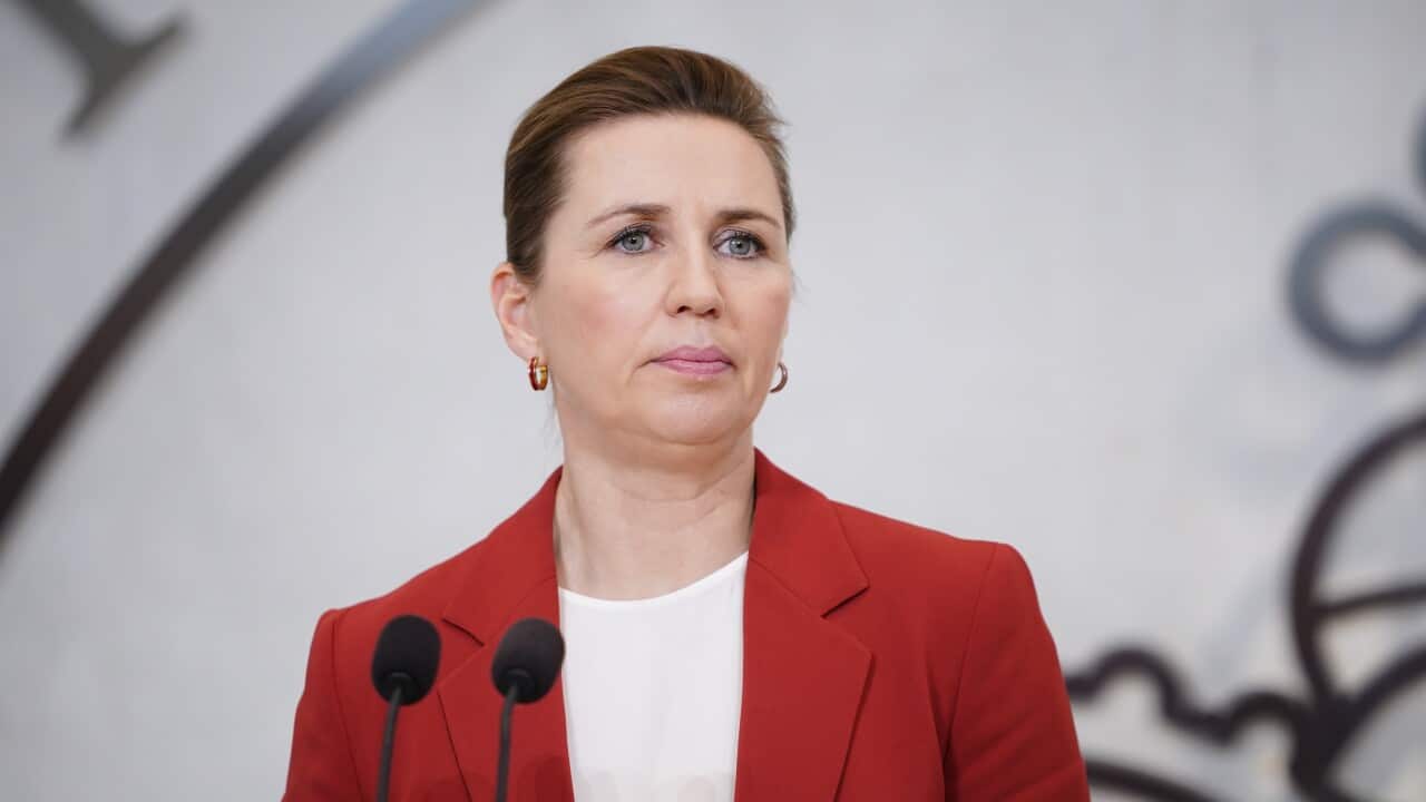 Danish Prime Minister Mette Frederiksen stands in front of two microphones wearing a red blazer.