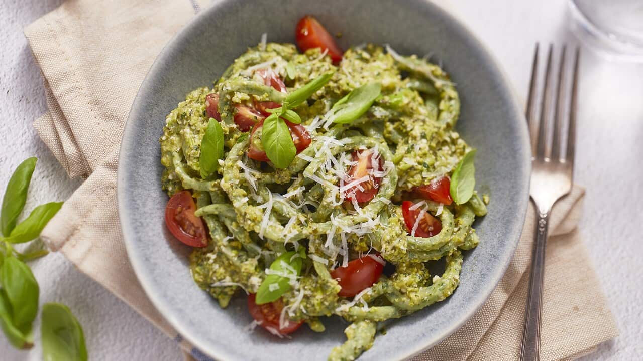 A pale speckled grey-blue bowl holds fat long pasta shapes, in a chunky green sauce, dotted with fresh cherry tomato pieces and scattered with grated parmesan.
