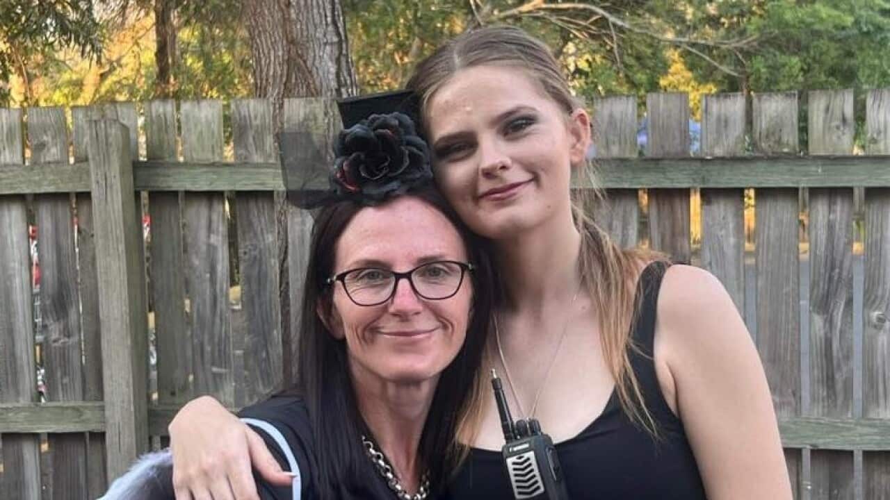 A young woman in a black top on the right is standing with her hand around the shoulder of a middle-aged woman also in a black outfit.