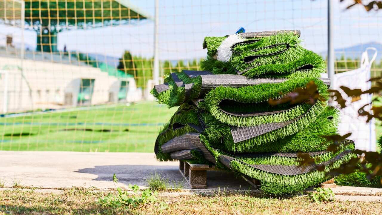 A pile of artificial turf is lying on a path next to a football field.