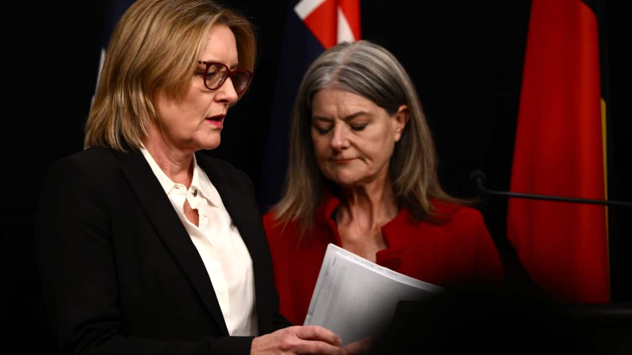 A woman in a black jacket and white shirt, holding a document, is standing next to a woman in a red jacket.