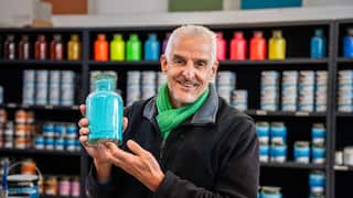 A man wearing a grey sweater stands holding a jar of blue paint powder, in front of shelves stacked with paint pots.