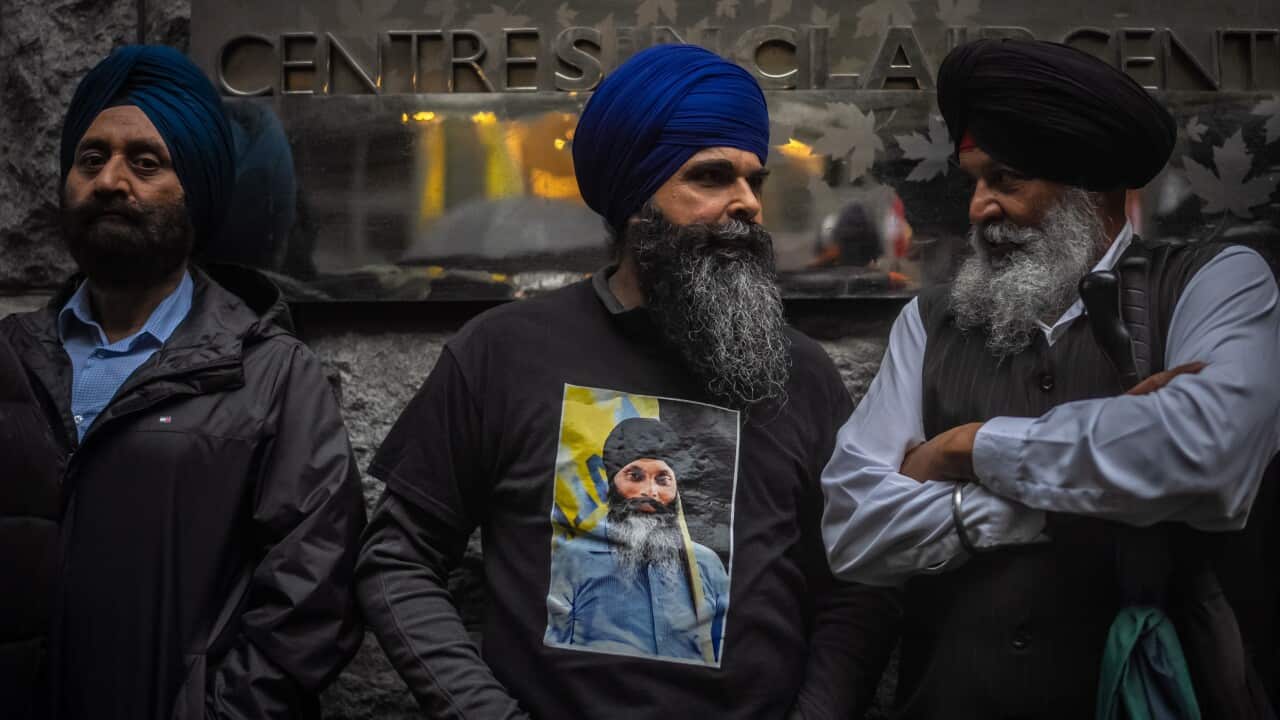 Three Sikh men are standing in protest, with the one in the middle displaying a poster of a Sikh man taped to his chest.