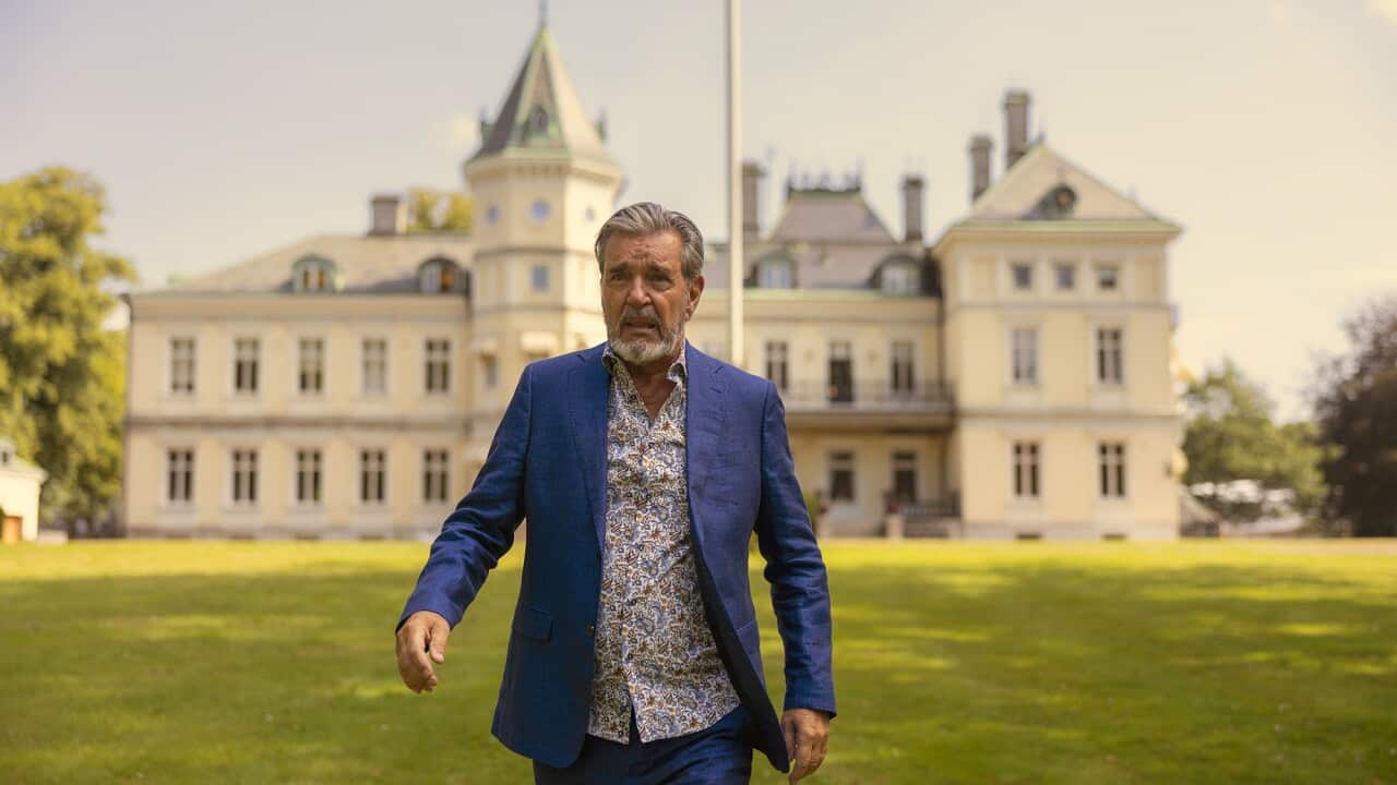 A grey-breaded man in a blue jacket walks away from an impossing multistory European building.