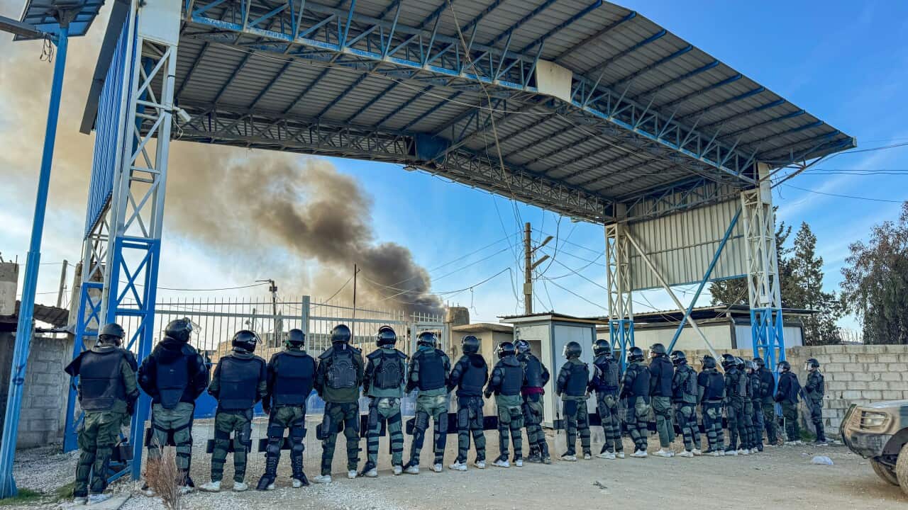 A group of riot police in dark uniforms, helmets, and protective gear stands in a line under a large metal canopy, facing a metal gate. In the background, a large plume of dark smoke rises into the blue sky.