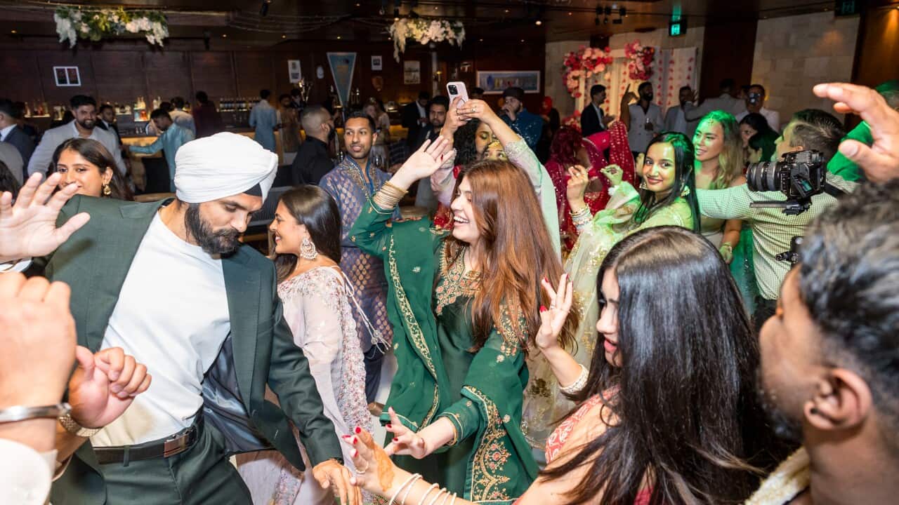 A group of people dressed in a mix of traditional and modern attire dances in a dimly lit, richly decorated indoor wedding reception setting.