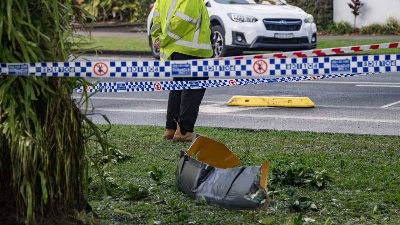 Police tape has cordoned off the crash site, which includes some debris. A worker wearing a yellow hi-vis and a police vehicle are visible in the background.