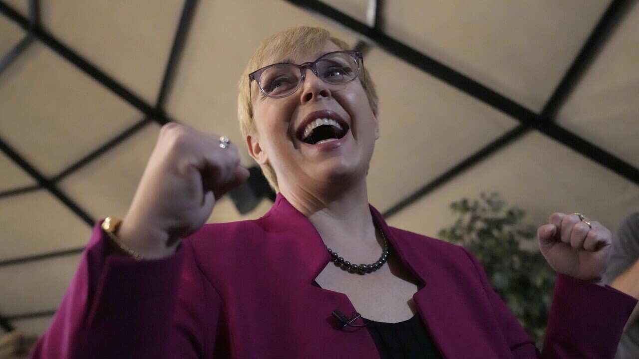 A blonde woman in a magenta jacket holds up two fists in celebration