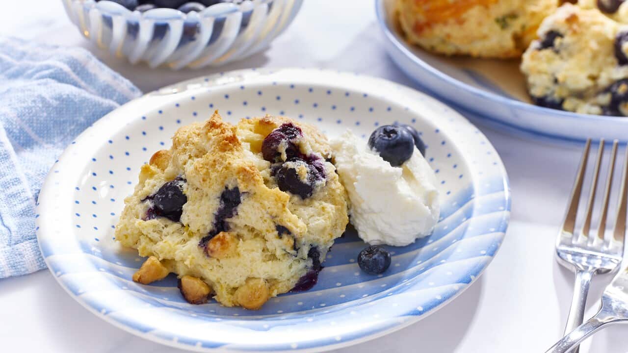 A shallow blue and white bowl with a spot and stripe pattern holds a rough-shaped baked scone-like biscuit. A scoop of cream and a few blueberries sit alongside, in the bowl. A tray can be seen in the background with more biscuits.