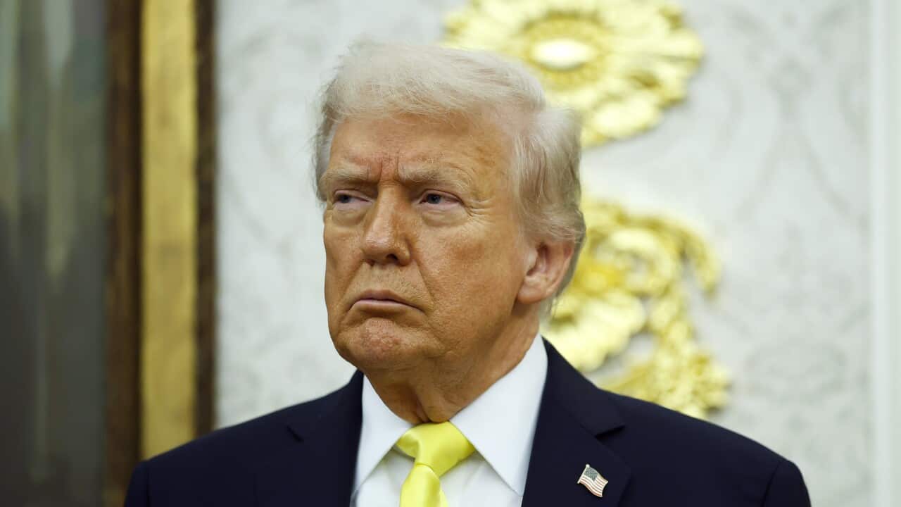Donald Trump frowns, wearing a black suit and a yellow tie. Behind him is an out-of-focus gold ornamental design on a wall.