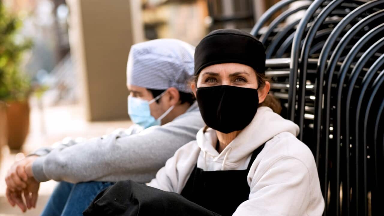Mature female cook posing with a male young coworker wearing both protective face masks sitting on the floor outside the restaurant they work