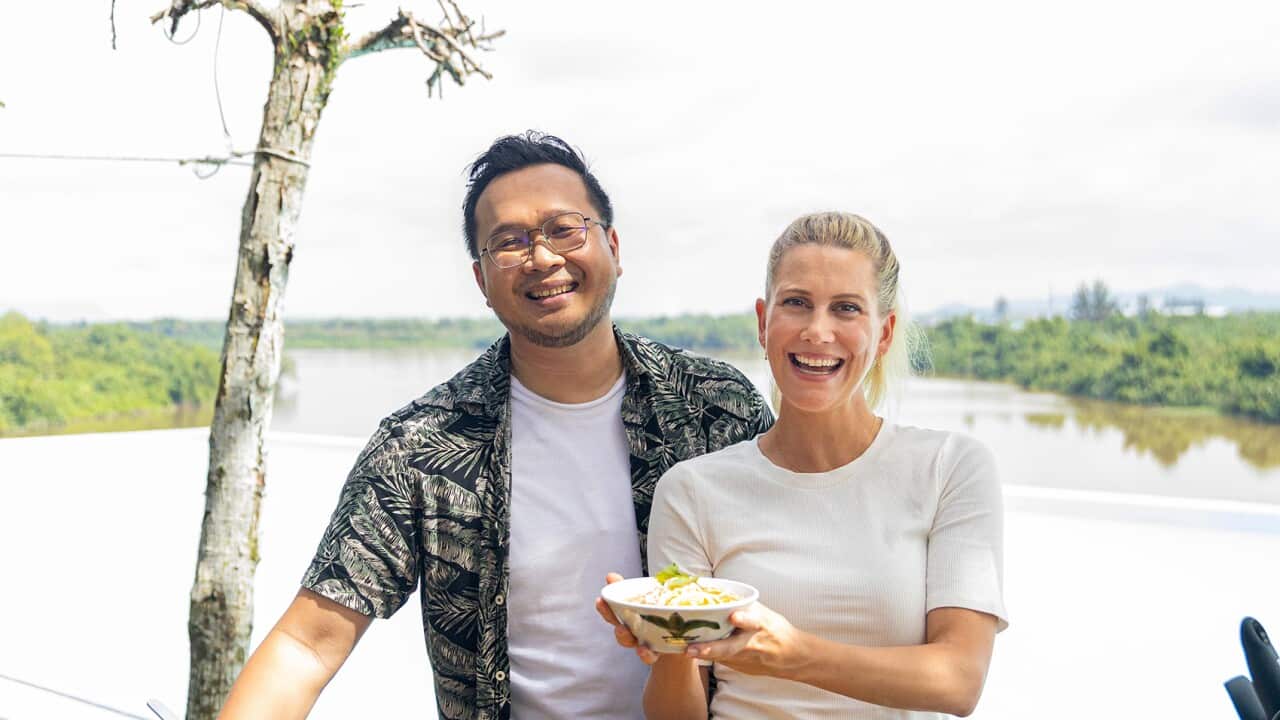A smiling Asian man and a blonde woman stand behind a table, which has various cooking items and food stuffs on it. The woman is holding a bowl of noodles. A wide body of water fills the background.