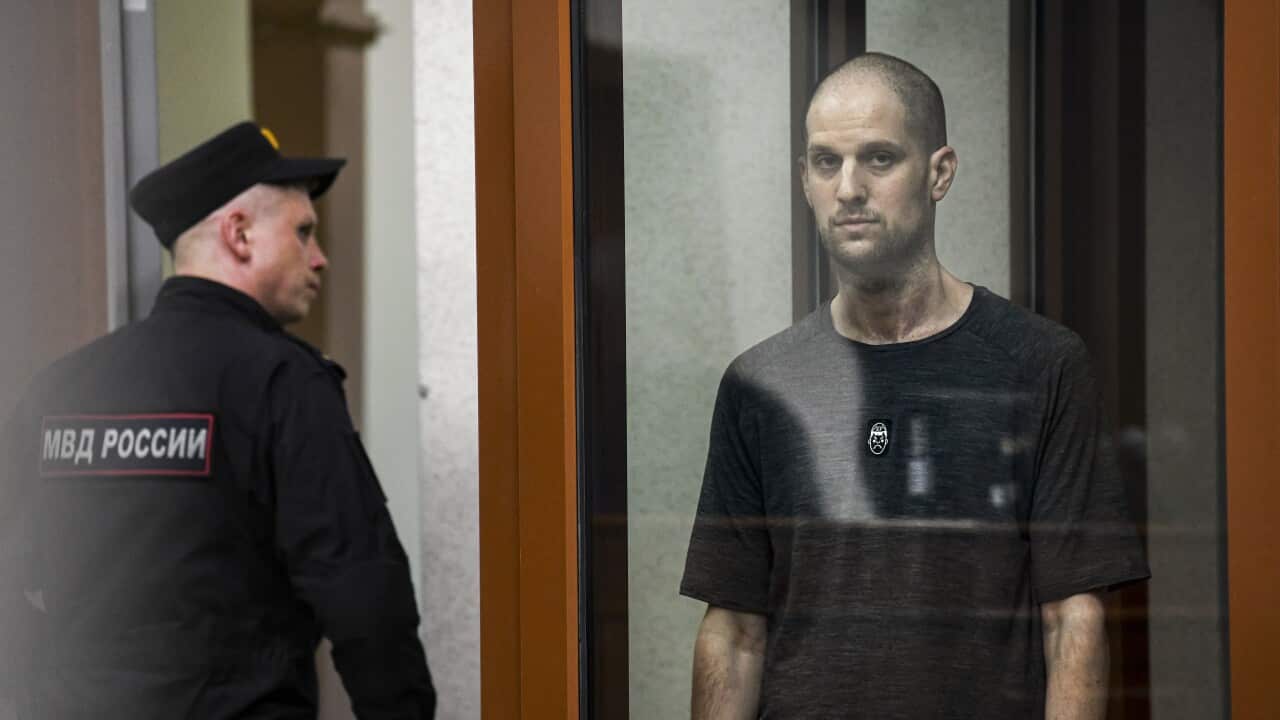 A man in a grey t-shirt standing in a glass cell in a courtroom