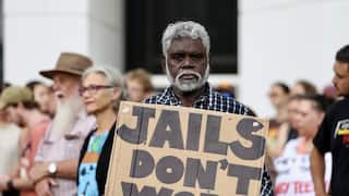 Man looks at the camera, holding a cardboard sign that reads "JAILS DON'T WORK" in front of a crowd of people walking.