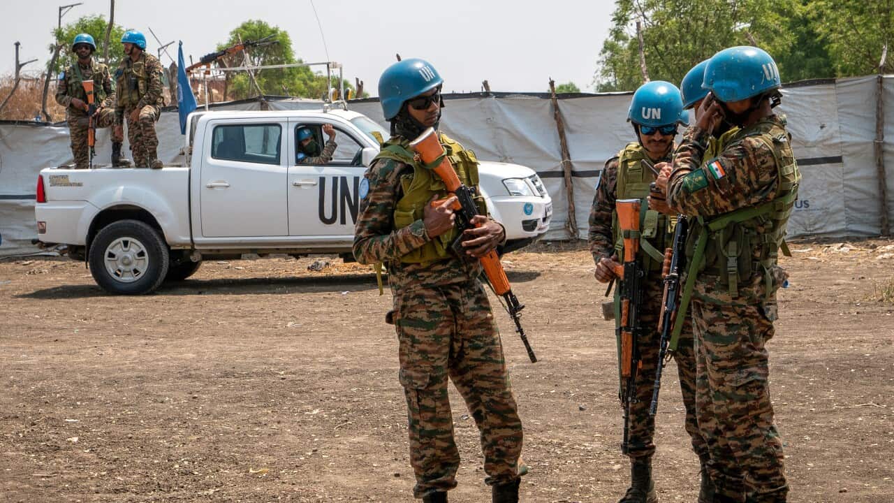 Three men holding firearms stand wearing camouflage and blue United Nations helmets in front of a white UN car