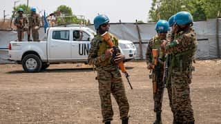 Three men holding firearms stand wearing camouflage and blue United Nations helmets in front of a white UN car