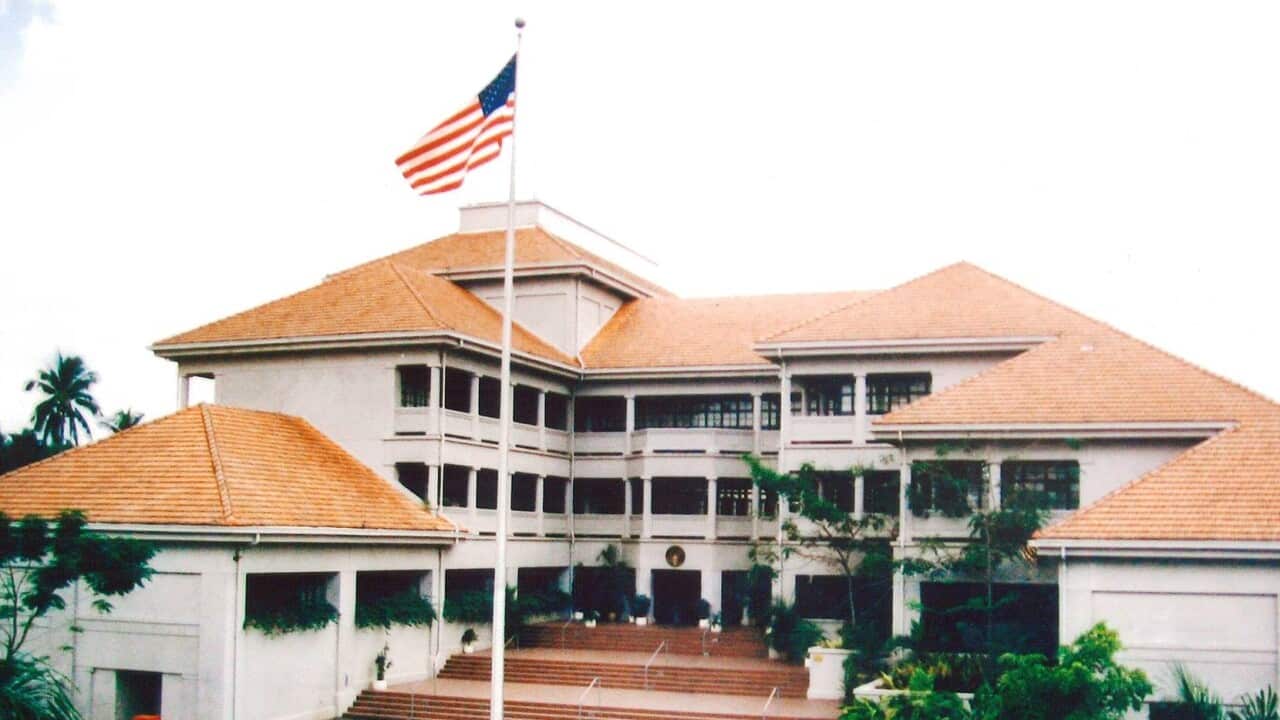 Donald Trump, wearing a suit and a red tie, stands in front of the US flag.