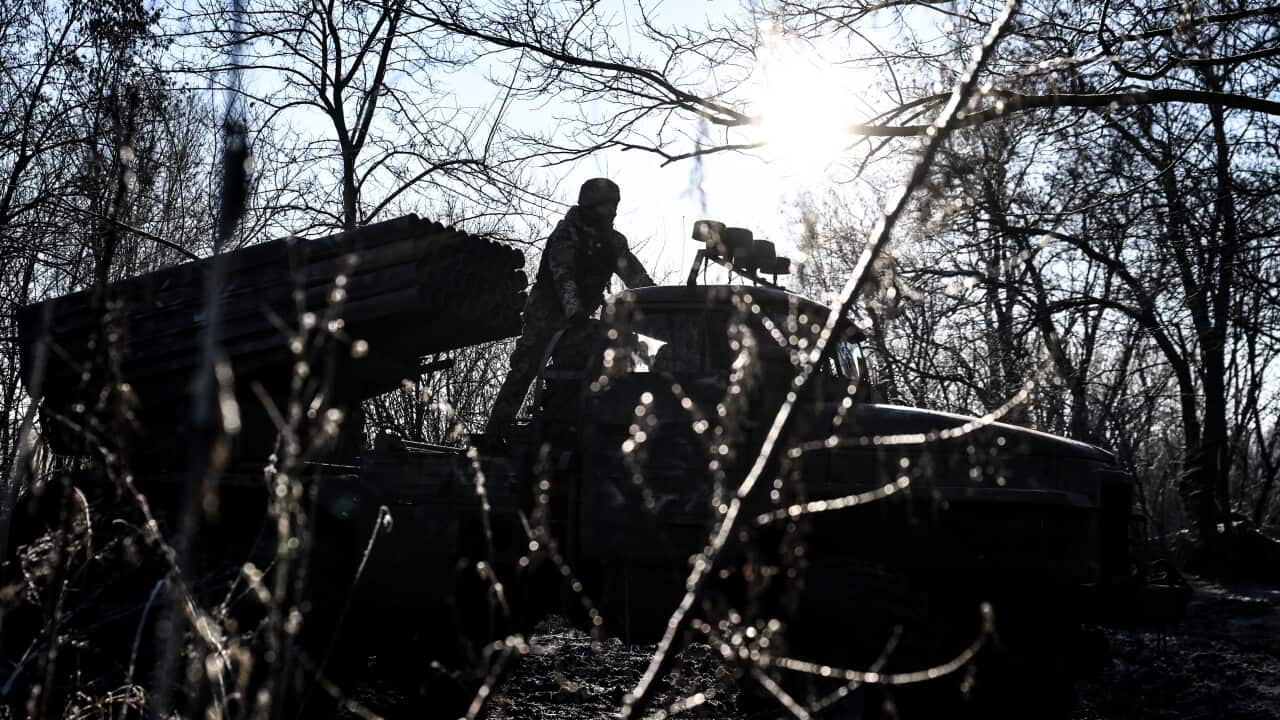 A soldier stands atop a truck-mounted multiple rocket launcher system positioned in a wooded area, silhouetted against a bright, low-hanging sun.