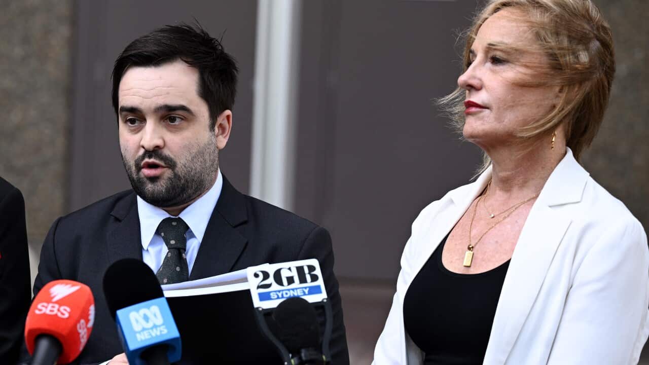 A man with a beard and black hair wearing a suit speaks before microphones as he stands beside a blonde woman.