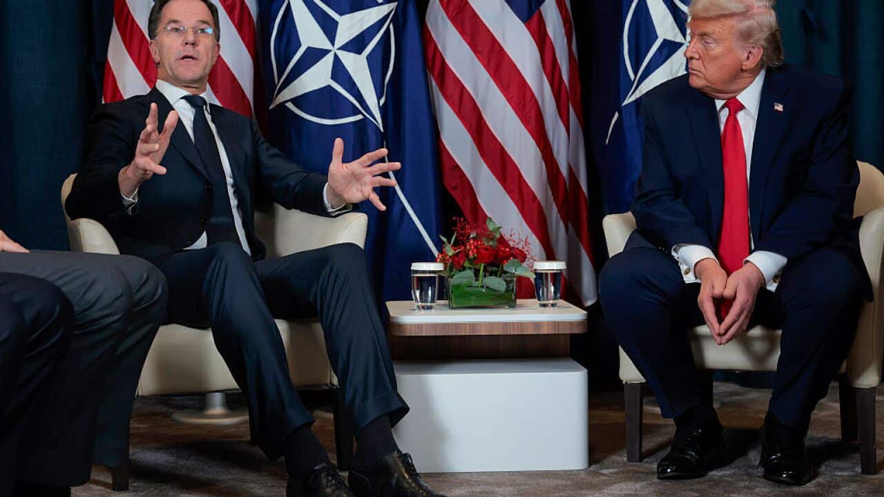 Two men in suits sit together in front of flags representing the United States and NATO.