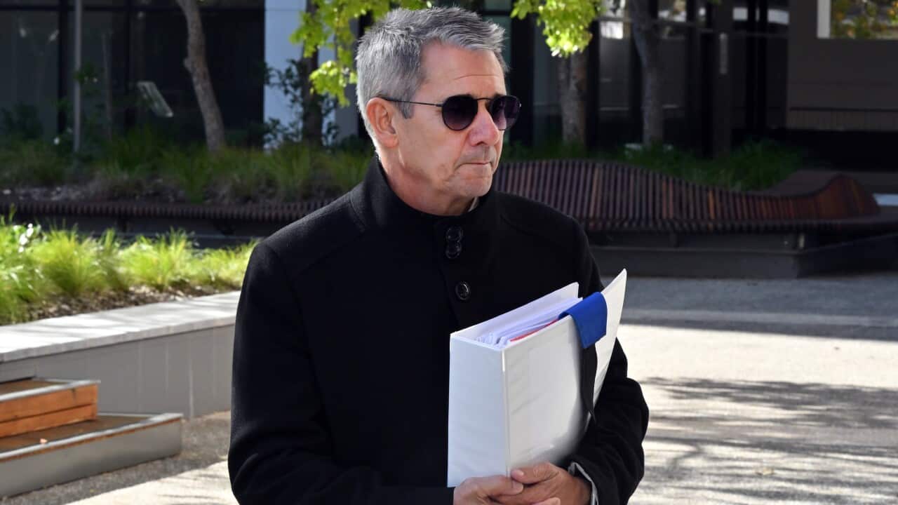 A man in a black jacket and sunglasses holds a large white folder.