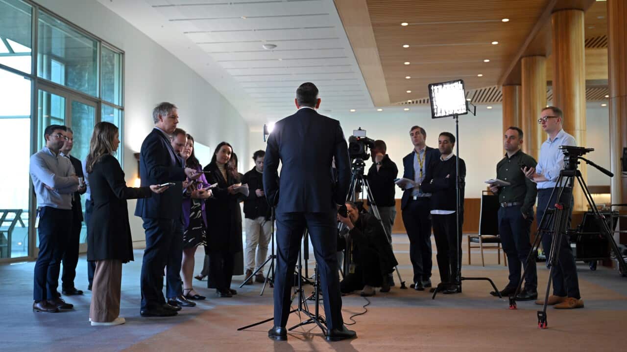 A man in a blue suit with his back turned in front of a row of people and cameras.