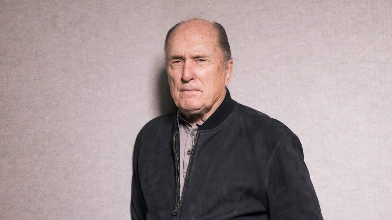 A man wearing a black jacket, standing in front of a grey wall.