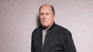 A man wearing a black jacket, standing in front of a grey wall.