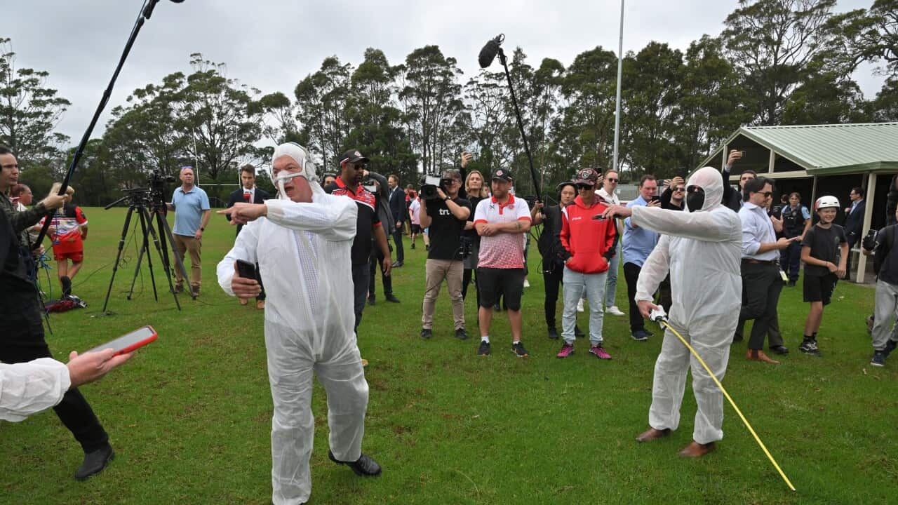Two men dressed in white, hazmat-like suits, surrounded by people holding cameras, outside.
