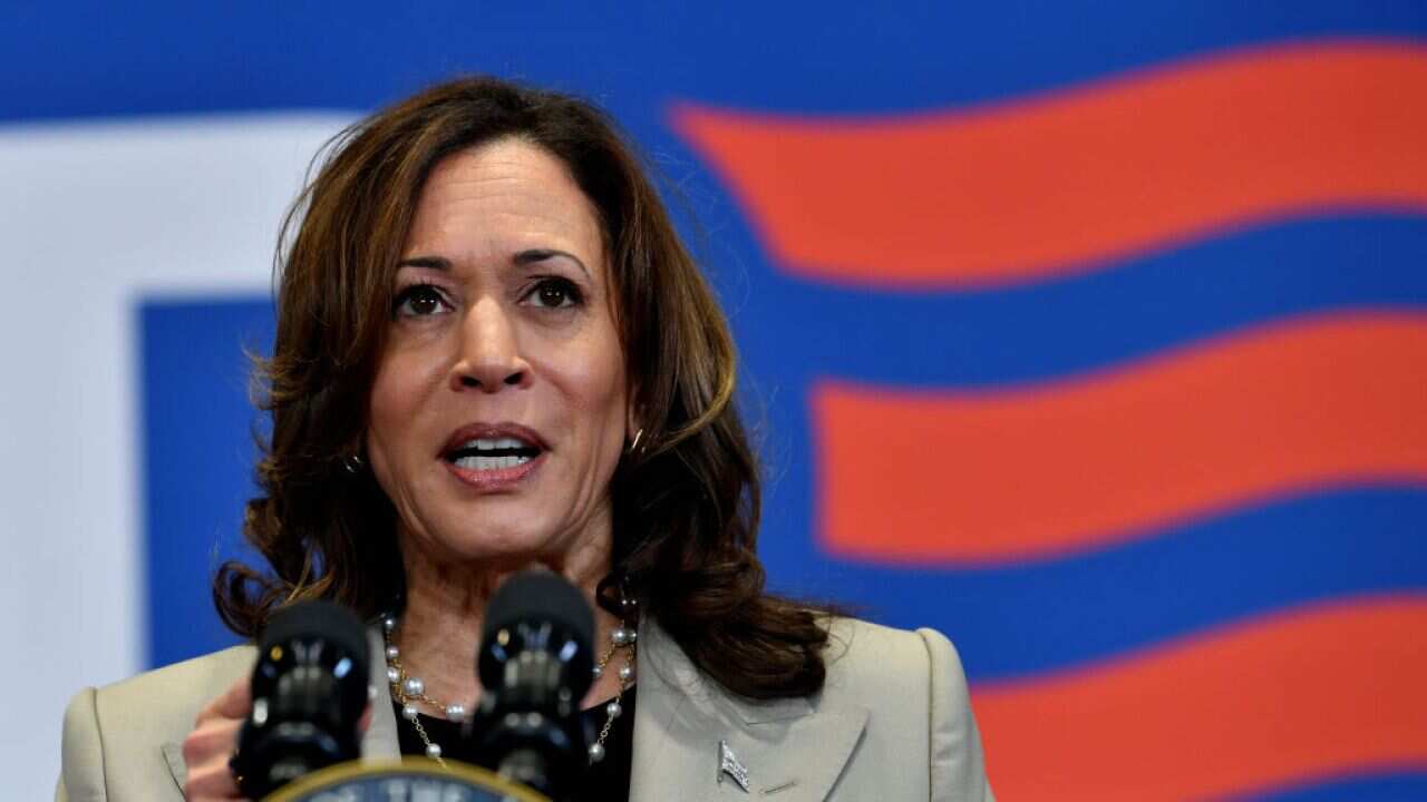 A woman stands at a podium with a microphone, with the US flag in the background.