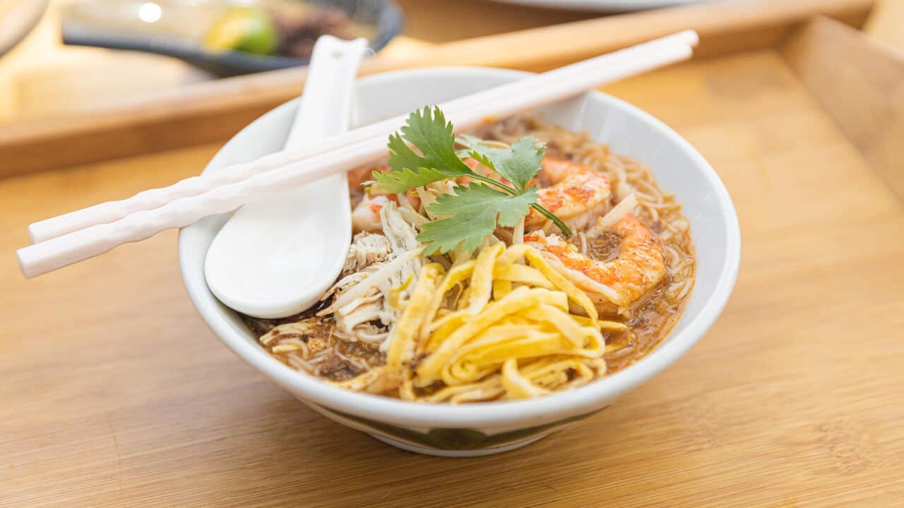 A white bowl sits on a wooden surface. It holds a laksa, with prawns visible.