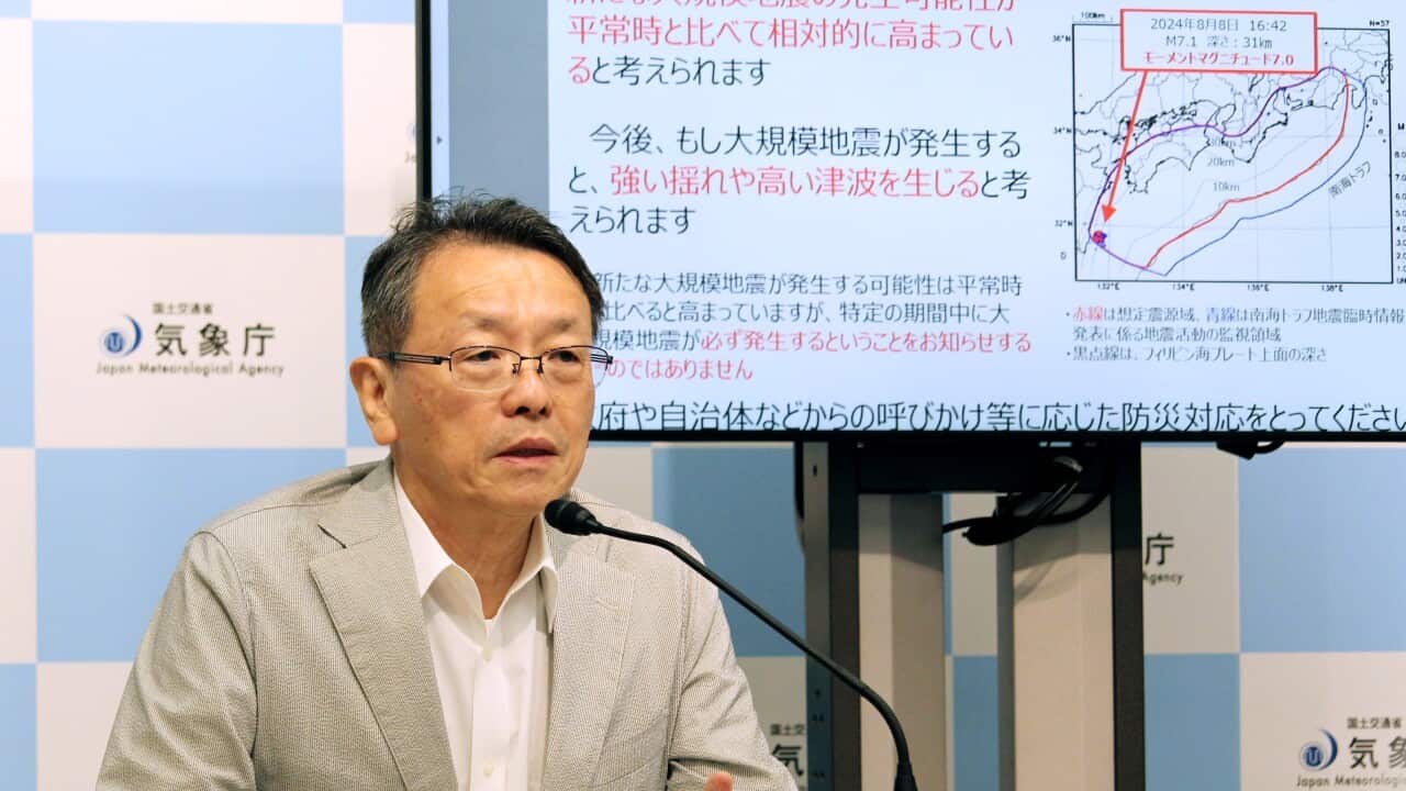 A man wearing glasses and a tan suit jacket speaks into a microphone. Behind him, a projected image of geological information and Japanese text is visible.