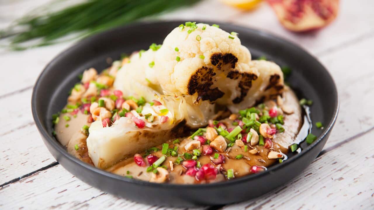 A cauliflower wedge with a darkened cut side sits in a shallow black bowl, on a pool of creamy sauce. The dish is decorated with chopped chives and pomengranate seeds