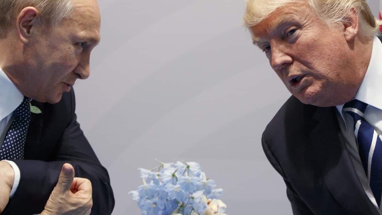 A man in a suit leans over to speak to another man in a suit with a flower vase between them