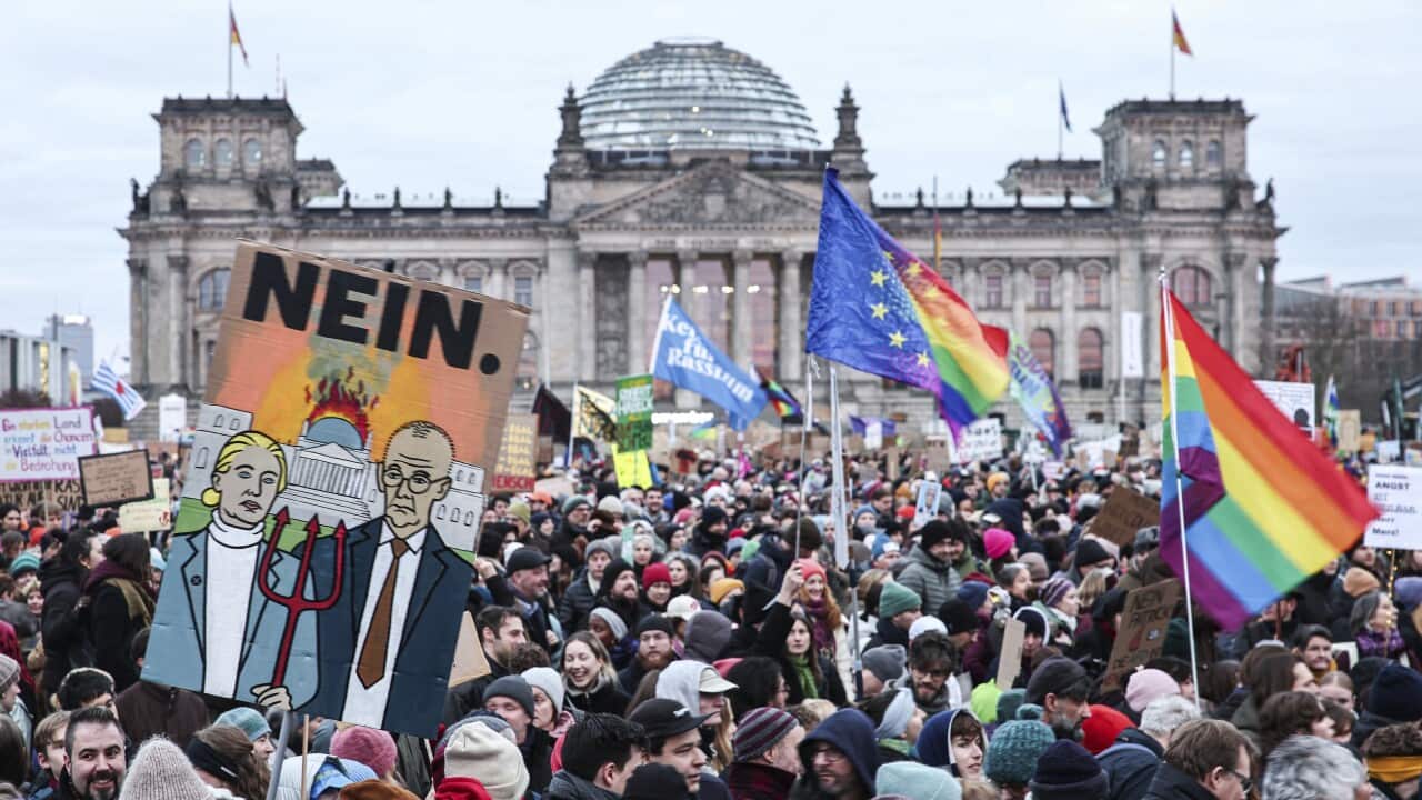 Thousands of protesters gather outside a government building, some holding flags and posters.