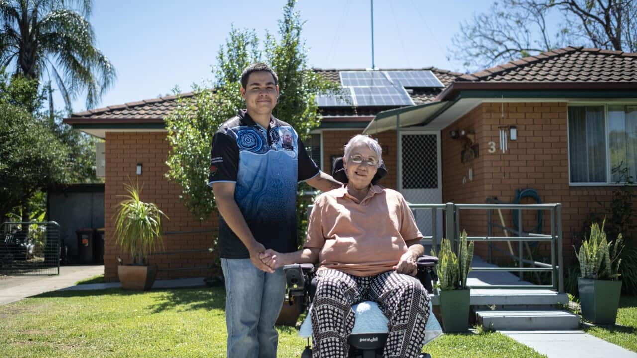 Image of a man wearing a blue tshirt and jeans with his arm wrapped around his grandma who is sitting in a motorised chair outside their suburban home