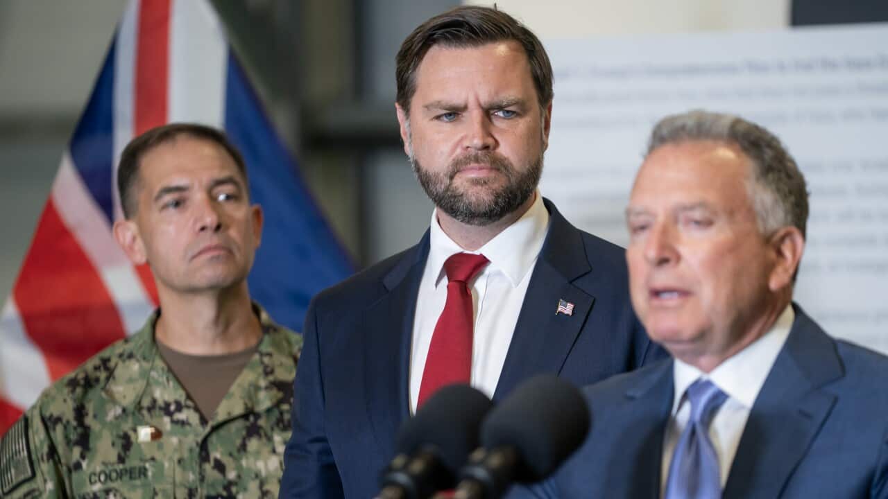 A man in a blue suit is speaking into a microphone, flanked by another man in a blue suit and a military official.