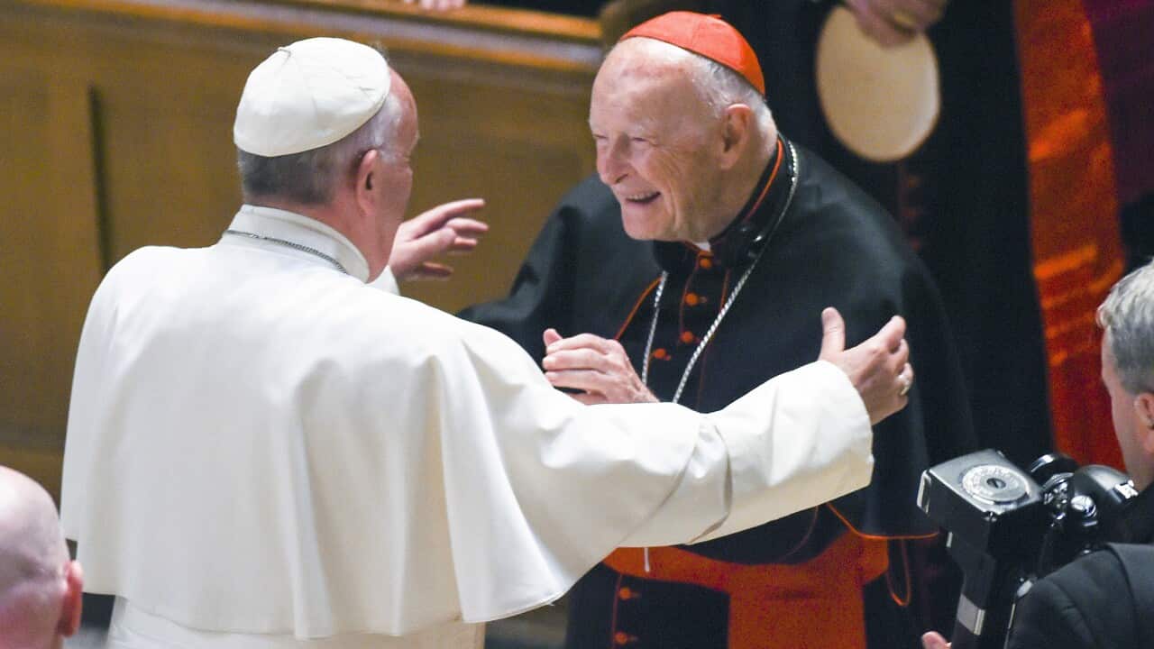 Pope Francis, dressed in white, reaches out to hug a man dressed in black cardinal robes with red trim